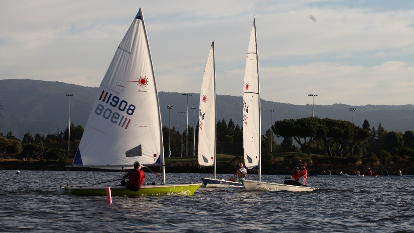 Three laser saiboats on shoreline lake.