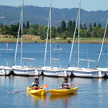 Two kids in kayaks on shoreline lake.